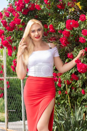 Téléchargez les photos : Belle femme aux cheveux blonds en haut blanc et longue jupe rose posant dans le jardin fleuri avec des roses rouges - en image libre de droit