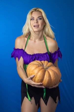 Photo for Young beautiful blonde woman in a witch costume on a blue background. Holds a large pumpkin in his hands. Short dress, long hair. Halloween concept. Soft focus - Royalty Free Image