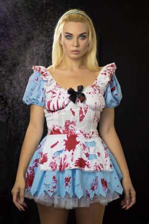 Téléchargez les photos : Belle jeune femme blonde en costume Alice. La robe est déchirée et tachée de faux sang. Concept d'Halloween. Concentration douce - en image libre de droit