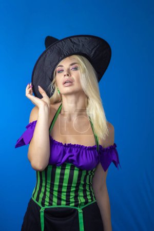 Photo for Young beautiful blonde woman in a witch costume on a blue background. Short dress, long hair, black hat. Halloween concept. Soft focus - Royalty Free Image