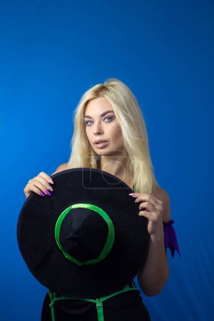 Photo for Young beautiful blonde woman in a witch costume on a blue background. Short dress, long hair, black hat. Halloween concept. Soft focus - Royalty Free Image