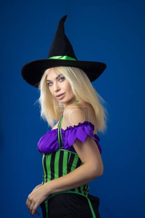 Téléchargez les photos : Jeune belle femme blonde en costume de sorcière sur fond bleu. Robe courte, cheveux longs, chapeau noir. Concept d'Halloween. Concentration douce - en image libre de droit