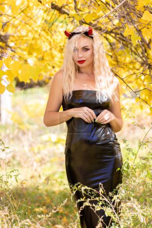 Téléchargez les photos : Belle femme blonde en costume de diable dans la nature. Robe serrée en latex noir et cornes. Concentrez-vous. Concept Halloween - en image libre de droit