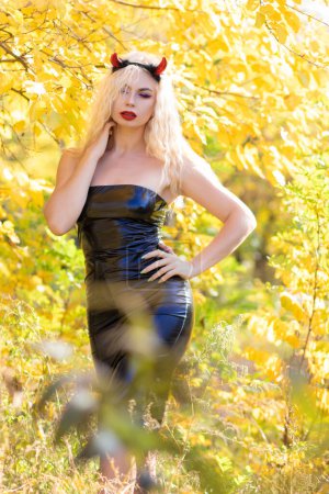 Photo for Beautiful blonde woman in a devil costume in nature. Black latex tight dress and horns. Soft focus. Halloween concept - Royalty Free Image