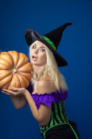 Photo for Young beautiful blonde woman in a witch costume on a blue background. Holds a large pumpkin in his hands. Short dress, long hair. Halloween concept. Soft focus - Royalty Free Image