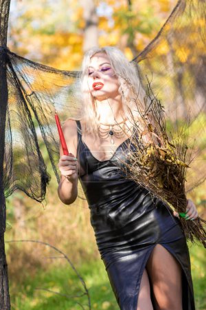 Foto de Sesión de fotos de una rubia con el pelo ondulado en la imagen de una bruja: un vestido de cuero largo y una escoba en las manos. Concepto Halloween - Imagen libre de derechos