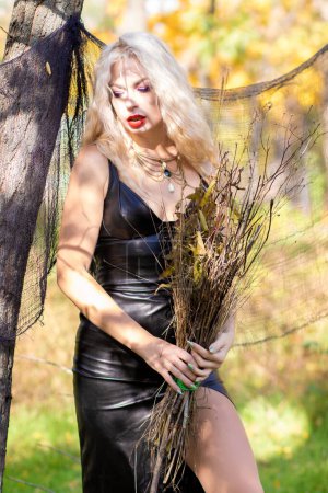 Photo for Photo shoot of a blonde with wavy hair in the image of a witch: a long leather dress and a broom in her hands. Halloween concept - Royalty Free Image