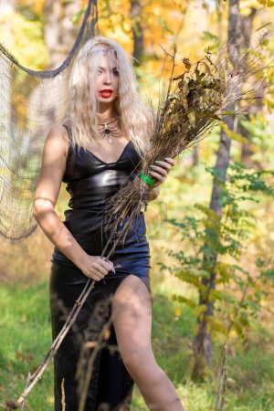 Foto de Sesión de fotos de una rubia con el pelo ondulado en la imagen de una bruja: un vestido de cuero largo y una escoba en las manos. Concepto Halloween - Imagen libre de derechos