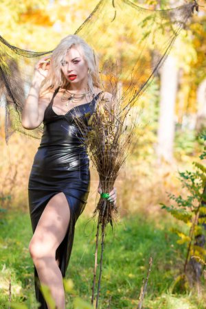Téléchargez les photos : Séance photo d'une blonde aux cheveux ondulés à l'image d'une sorcière : une longue robe en cuir et un balai à la main. Concept Halloween - en image libre de droit