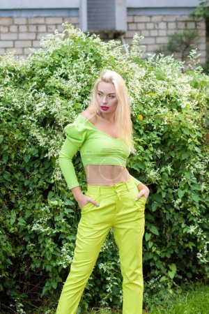 Foto de Retrato de larga duración de una hermosa rubia joven con el pelo largo en un traje de pantalón amarillo-verde y brillantes bombas de tacón alto sobre un fondo de arbustos verdes con flores - Imagen libre de derechos
