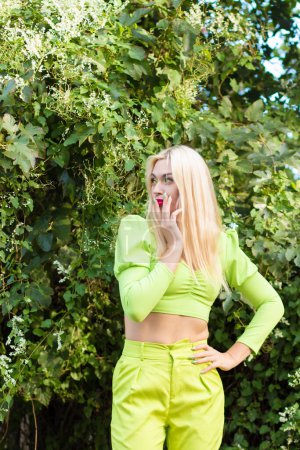 Foto de Retrato de una hermosa rubia joven con el pelo largo en un pantalón amarillo-verde sobre un fondo de arbustos verdes con flores - Imagen libre de derechos