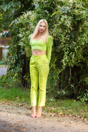 Téléchargez les photos : Portrait complet d'une belle jeune blonde aux cheveux longs en pantalon jaune-vert et aux talons hauts brillants sur fond de buissons verts fleuris - en image libre de droit
