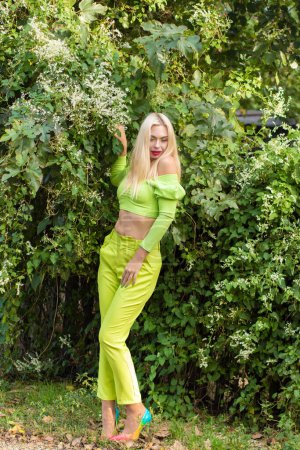 Téléchargez les photos : Portrait complet d'une belle jeune blonde aux cheveux longs en pantalon jaune-vert et aux talons hauts brillants sur fond de buissons verts fleuris - en image libre de droit