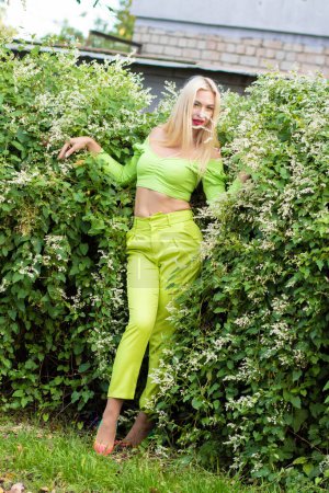 Photo for Full-length portrait of a beautiful young blonde with long hair in a yellow-green pantsuit and bright high-heeled pumps against a background of green bushes with flowers - Royalty Free Image