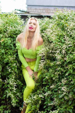 Foto de Retrato de larga duración de una hermosa rubia joven con el pelo largo en un traje de pantalón amarillo-verde y brillantes bombas de tacón alto sobre un fondo de arbustos verdes con flores - Imagen libre de derechos