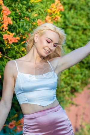 Téléchargez les photos : Portrait d'une femme blonde aux cheveux longs portant un haut blanc satiné et une jupe rose satiné. Coucher de soleil. Près des buissons avec de grandes fleurs orange - en image libre de droit