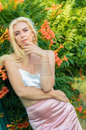 Téléchargez les photos : Portrait d'une femme blonde aux cheveux longs portant un haut blanc satiné et une jupe rose satiné. Coucher de soleil. Près d'une clôture avec des buissons aux grandes fleurs orange - en image libre de droit