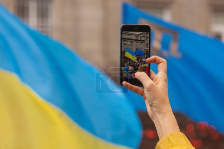 Photo for Ukrainians and supporters rally concept. The first anniversary of Russia's invasion of Ukraine. Hand shooting meeting on mobile phone. Close up. Copy space. Outdoor shot - Royalty Free Image