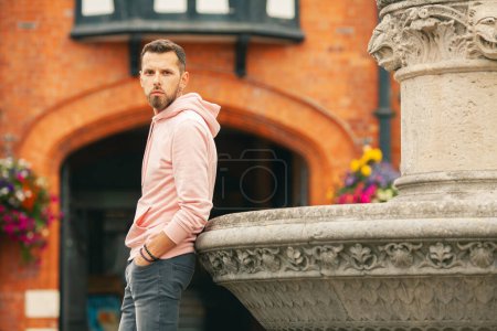 Photo for Young bearded romantic man wearing pink trendy hoodie, gray jeans posing over brick house with flowers on backgound. Old classical fontain. Outdoor shot - Royalty Free Image
