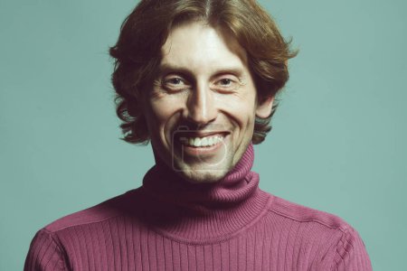 Photo for Male beauty concept. Emotional portrait of laughing handsome man in violet knitted polo neck posing over light blue background. Glossy long hair, shiny perfect smile. Studio shot - Royalty Free Image