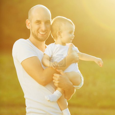 Téléchargez les photos : Concept de père et fils. Père souriant et son petit garçon portant des t-shirts blancs et posant ensemble. Journée ensoleillée d'été. Rayons de lumière. Plan extérieur - en image libre de droit