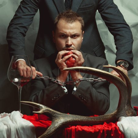 Photo for Devil concept. Portrait of captured criminal eating fresh heart. Policeman holding hands of cannibal with metal chain. Decadent accessories. Cinematic style. Studio shot - Royalty Free Image