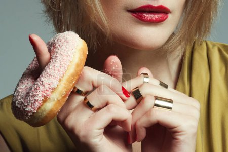 Photo for Dominant diva concept. Luxurious young model in mustard dress eating pink donut over gray background. Perfect blond hair, skin, make-up, manicure. Golden rings on fingers. Close up studio shot - Royalty Free Image