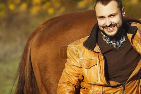 Foto de Concepto de vida rural. Joven hombre rico relajado y sonriente en chaqueta de cuero amarillo de pie cerca de caballo. Estilo vaquero retro. De cerca. Espacio de texto. tiro al aire libre - Imagen libre de derechos