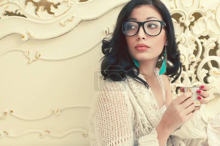 Photo for Cosy morning concept. Portrait of young gorgeous brunette having breakfast - tea (coffee) and posing in vintage bed. Perfect make-up and manicure. Trendy eyewear. Retro style. Indoor shot - Royalty Free Image