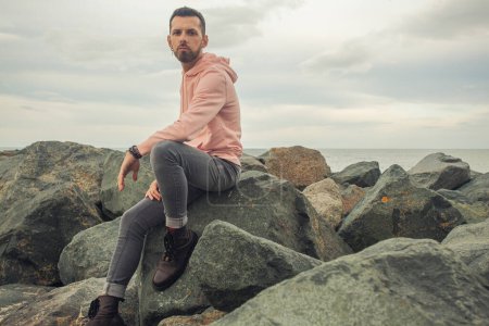 Foto de Concepto de moda masculino. Joven hombre guapo barbudo sentado en ropa de moda en la costa del mar pedregoso. Estilo callejero. Clima nublado. Espacio de texto. tiro al aire libre - Imagen libre de derechos