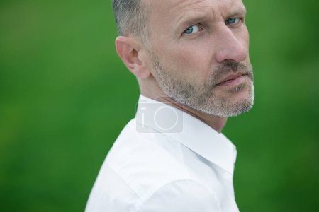 Photo for Fabulous at any age, male fashion concept. Portrait of 45-year-old man with blue eyes standing over green grass background in white shirt. Close up. Classic style. Gray hair. Outdoor shot - Royalty Free Image