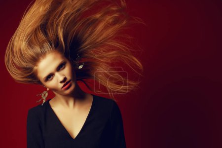 Téléchargez les photos : Concept ultra mode. Portrait de belle fille aux cheveux roux avec des cheveux volants porter robe de cocktail noire, posant sur fond rouge. Espace de copie. Plan studio - en image libre de droit