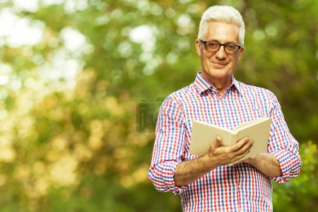 Photo for Active funny grandpa concept. Portrait of smiling handsome old man wearing stylish eyeglasses walking in the park and reading book. Fashionable casual clothing. Copy-space. Outdoor shot - Royalty Free Image
