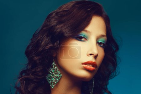 Photo for Epoque of Disco concept. Close up portrait of fashion model with curly hair and arty make-up posing over blue background. Vintage turquoise earrings. Perfect skin. Retro style. Copy-space. Studio shot - Royalty Free Image
