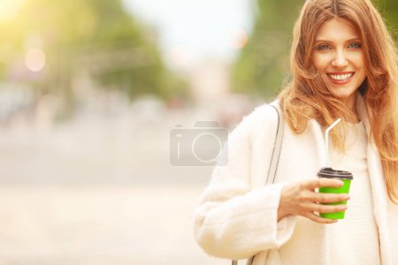 Photo for Coffee lover concept. Luxurious smiling woman drinking hot beverage through sipper at street of European city. Wind blown long hair. Model wearing white coat. Copy-space. Outdoor shot - Royalty Free Image