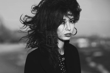 Photo for High fashion concept. Gorgeous young girl with long dark glossy hair in black dress and coat on the beach. Cinema star, diva style. Close up. Text space. Monochrome outdoor shot - Royalty Free Image