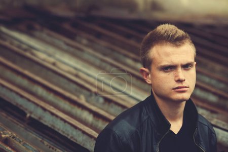 Photo for Bad boy concept. Portrait of brutal young man with short hair wearing black leather jacket, posing over urban corrugated background. Text space. Close up. Outdoor shot - Royalty Free Image