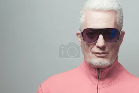 Foto de Fabuloso a cualquier edad, concepto de gafas. Primer plano retrato de un hombre de 60 años de moda con chaqueta rosa sobre fondo gris. corte de pelo de moda, pelo gris brillante. Estilo Fashionista. Captura de estudio - Imagen libre de derechos