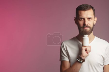 Téléchargez les photos : Super concept de supplément. Portrait d'un jeune beau mec actif avec une bouteille en plastique blanc posant sur fond rose et lavande. Espace de copie. Plan studio - en image libre de droit