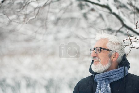 Téléchargez les photos : Concept de grand-père actif. Portrait d'un bel homme mature vêtu de vêtements d'hiver tendance et de lunettes à la mode debout sous un arbre, sur fond de parc à neige. Ferme là. Espace texte. Plan extérieur - en image libre de droit