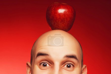Photo for Super Idea concept. Portrait of a funny scared and confused man with fearful glance posing over red background with red apple on head. Close up. Text space. Studio shot - Royalty Free Image