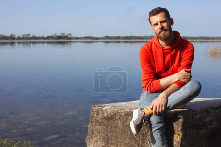 Photo for Discovering Ireland concept. Portrait of a young handsome man in red hoodie against the backdrop Lough Ennell in Belvedere House near Mullingar, County Westmeath. Outdoor shot - Royalty Free Image