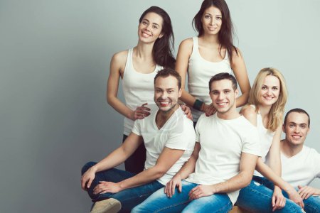Photo for Happy together concept. Group portrait of healthy boys & girls in white t-shirts, sleeveless shirts and blue jeans standing, sitting, posing over gray background. Urban street style. Copy-space. Studio shot - Royalty Free Image