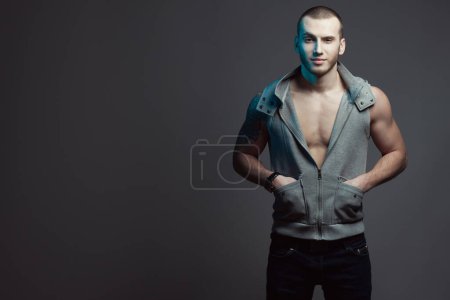 Photo for Portrait of smiling tattooed brutal young man with short hair & bristle on face wearing sleeveless jacket, blue jeans, posing over gray background. Hands in pockets. Copy-space. Studio shot - Royalty Free Image
