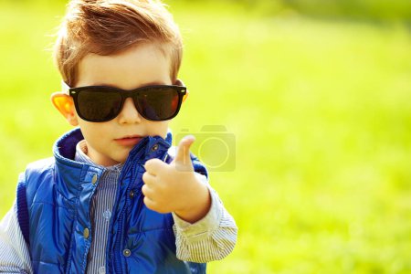 Photo for Stylish baby boy with ginger hair in trendy sunglasses and blue jacket standing in the park showing thumb up, ok sign. Hipster style. Sunny weather. Copy-space. Close up. Outdoor shot - Royalty Free Image