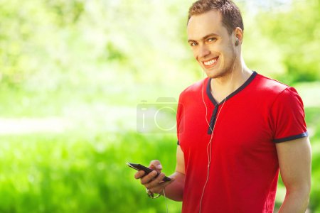 Gadget lover concept. Portrait of a young muscular man in red t-shirt and jeans walking with cell phone in the park. White shiny smile. Sunny weather. Close up. Copy-space. Outdoor shot