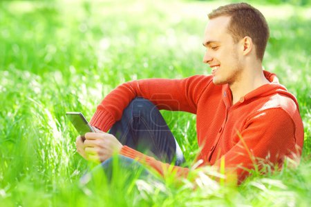 Gadget lover concept. Portrait of smiling young man in casual clothing reading e-book, sitting in green grass in the park. Great white shiny smile. Close up. Copy-space. Sunny weather. Outdoor shot