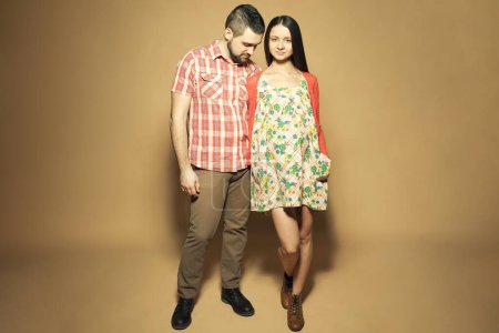 Photo for Stylish pregnancy concept. Full length portrait of good-looking family in trendy clothes (shirt, dress, cardigan, jeans and leather boots) expecting their child. Pastel colors. Copy-space. Studio shot - Royalty Free Image