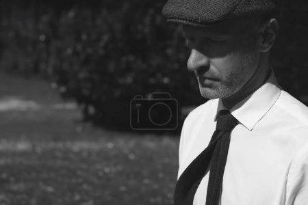 Photo for Mens fashion concept. Portrait of handsome mature man wearing white classic shirt, stylish tie and gray trendy cap walking in city park. Close up. Text space. Old films style. Outdoor monochrome shot - Royalty Free Image