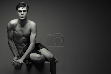Photo for Handsome muscular male model with nice abs posing undressed. Young man sitting on wooden cube. High fashion style. Copy-space. Black and white (monochrome) studio shot - Royalty Free Image